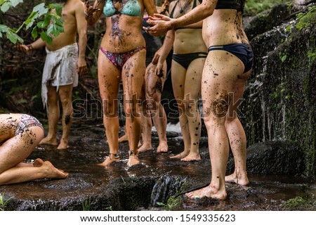 A mixed group of people are seen standing in a muddy stream with sludge covered legs and bodies during a sacred retreat in nature.