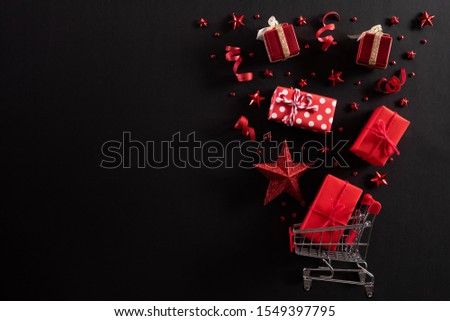 Christmas and Boxing Day background concept. Top view of shopping cart splash out Christmas decoration with gift box red star berries and ribbon on black background. Royalty-Free Stock Photo #1549397795