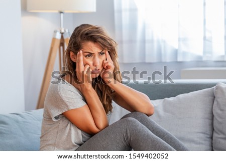 Portrait of an attractive woman sitting on a sofa at home with a headache, feeling pain and with an expression of being unwell.