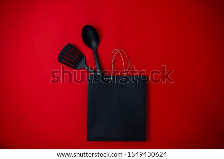 Black friday image with space for text. Bkack friday sale flat lay. Banner for home goods shop. Spatula, big spoon and black bag on the red background.