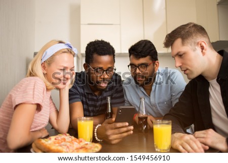 Four friends sitting at table, eating pizza, drinking soft beverages, reading text message on smartphone, testing new phone application together. Multicultural students having fun in hostel kitchen.