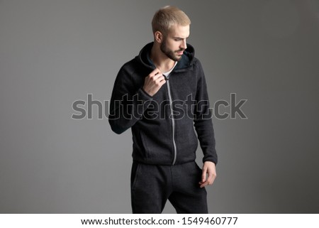 young casual man standing and fixing his tracksuit while looking aside pensive on gray studio background Royalty-Free Stock Photo #1549460777