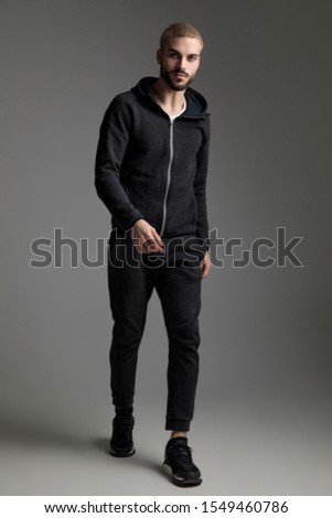 attractive casual man wearing tracksuit walking and staring at camera on gray studio background Royalty-Free Stock Photo #1549460786