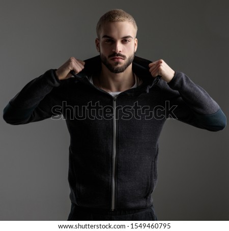beautiful casual man wearing tracksuit standing and fixing his hood while staring at camera confident on gray studio background Royalty-Free Stock Photo #1549460795