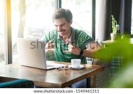 Yes. Young happy excited businessman in green t-shirt sitting, working and looking at laptop screen with rock sign and cheering. business and freelancing concept. indoor shot near window at daytime.