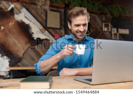 Joyful casual man reading, laughing, and drinking his coffee while wearing a blue shirt, sitting at a desk on coffeeshop background