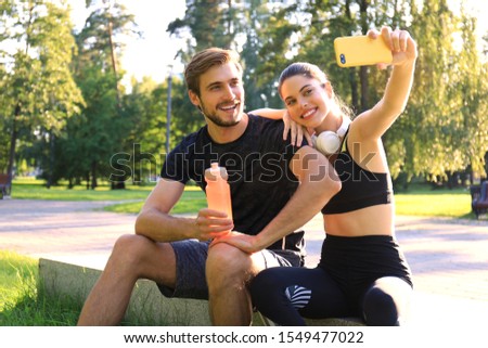 Young sporty couple in sportswear sitting on parapet in urban park and taking selfie after workout on summer sunny day.