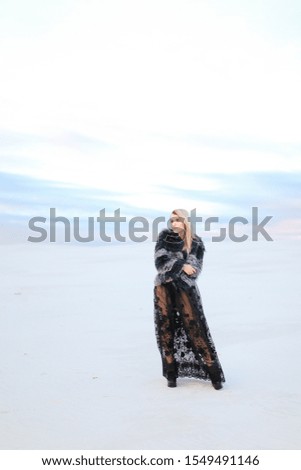 Young fashionable woman wearing black dress standing in white snowed steppe background. Concept of fashion and winter photo session.