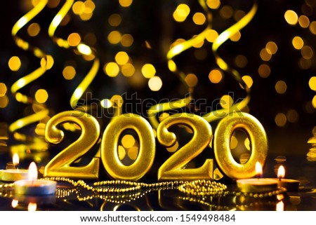Gold inscription 2020, burning candles, decor are on table. Defocused gold serpentine, festive decorative garland with yellow light bulbs, lanterns are shining on background. New year, christmas mood.