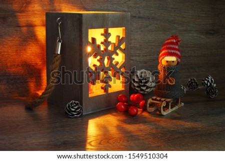 A wooden lantern and a figure of a boy in a gray sweater on a toy sled on a wooden background. Close up. Сandle light. Christmas still life. Scandinavian style. The negative space at the bottom.