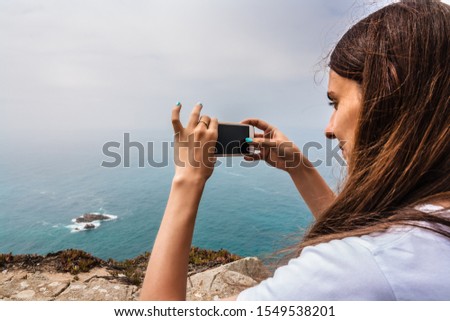 young woman photographs on a smartphone the landscape of the ocean and rocks from Cape Roca in Sintra, Portugal.