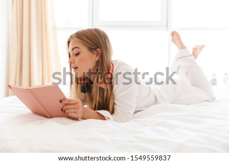 Photo of young caucasian woman in pajama with headphones making notes while lying on bed in light room