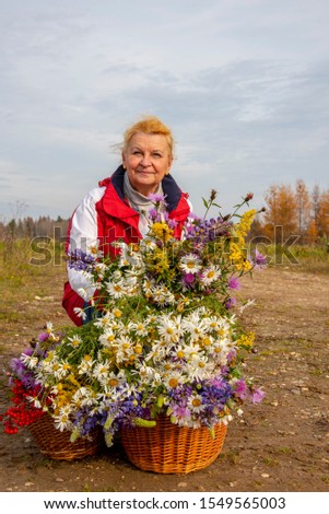 An aged woman sits on the road in a field with two baskets full of meadow bright flowers.