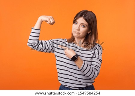 Portrait of proud confident woman with brown hair in long sleeve striped shirt raising arm pointng at biceps, boasting of female power, feminism. indoor studio shot isolated on orange background