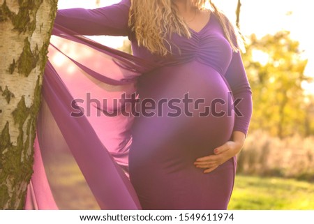 Portrait of beautiful pregnant woman in a purple dress, standing outside in beautiful autumn sunlight, stock picture.