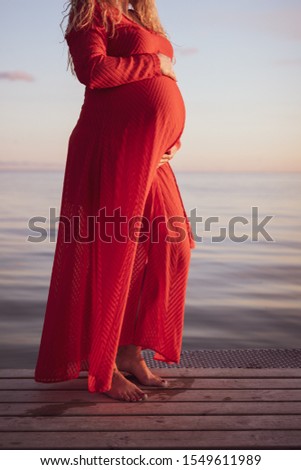 Portrait of beautiful pregnant woman in a red dress touching her stomach, standing outside by the ocean in beautiful autumn evening sunlight, stock picture.