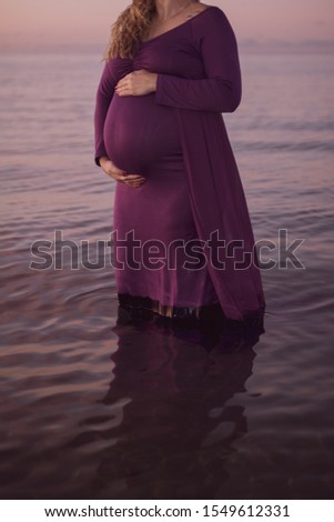 Portrait of beautiful pregnant woman in a purple dress touching her stomach, standing outside in the ocean in beautiful autumn evening sunlight, stock picture.