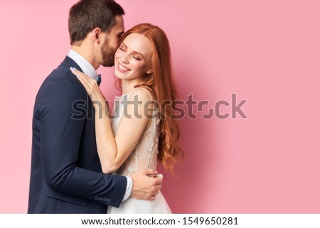 Caucasian man in suit and woman in white wedding dress with long hair lovely hug each other and smile, after wedding ceremony posing isolated over pink background.