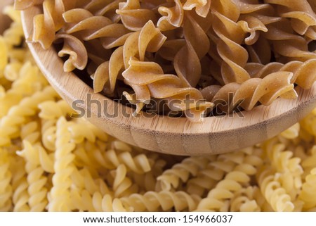 Raw food composition - yellow and brown rotini in a wooden bowl placed on a bright background.