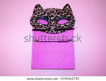 A pink diary and notebook with a cute masquerade cat mask in leopard pattern and a pen on. Pink background. Flat lay photography. 