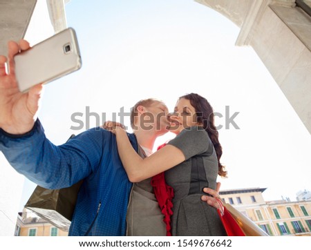 Beautiful consumer tourist couple kissing hugging on sunny city break holiday sightseeing together, using technology smartphone, taking selfies outdoors. Travel vacation, leisure recreation lifestyle.