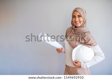 Young Muslim architect-woman wearing a protective helmet standing on the grey background. Pretty arabic architect woman. Ingenieur Arabic muslim woman with hijab holding protective helmet.