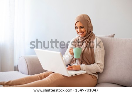 Young asian muslim woman sitting alone in a cafe working on her laptop and holding a coffee mug. Young muslim woman using white laptop. Beautiful young muslim woman using laptop at home
