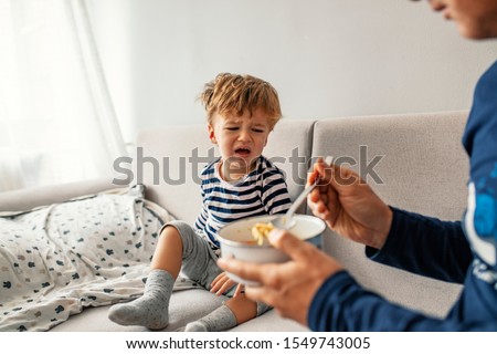 Unhappy child refusing to eat at home with white background while father is trying to deceive the toddler by tricks. Upset child refuse to eat healthy food and demanding bad unhealthy fastfood meals