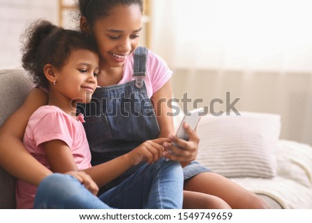 New generation addicted with gadgets concept. African teen girl teaching her baby sister to use mobile phone, free space