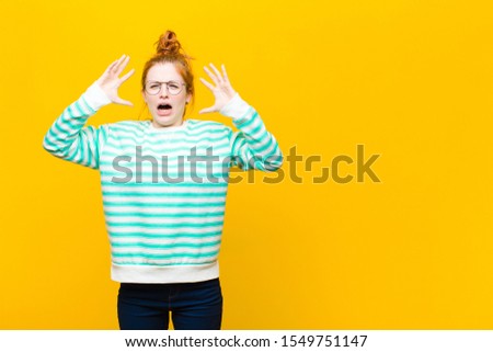 young red head woman screaming with hands up in the air, feeling furious, frustrated, stressed and upset against orange wall