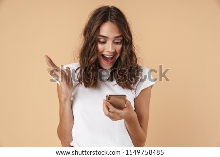 Portrait of a lovely cheerful young girl wearing casual clothing standing isolated over beige background, celebrating while holding mobile phone