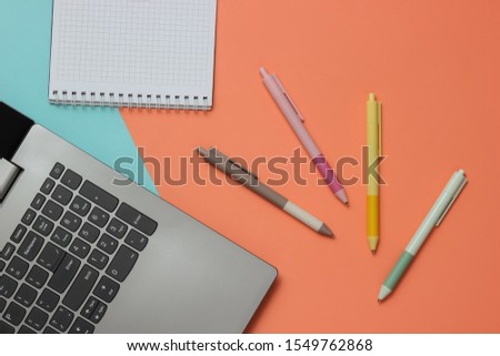 Laptop and many colored pens with notebook close-up on blue pink background. Top view