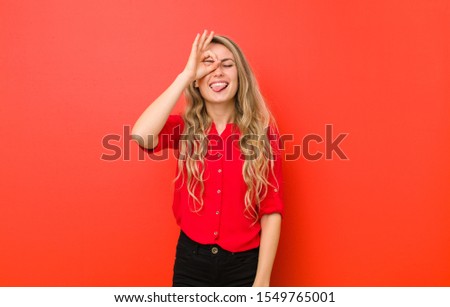 young blonde woman smiling happily with funny face, joking and looking through peephole, spying on secrets against red wall
