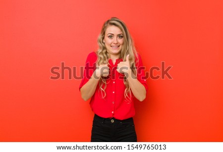 young blonde woman feeling happy, surprised and proud, pointing to self with an excited, amazed look against red wall