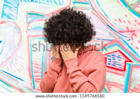 young pretty afro woman covering eyes with hands with a sad, frustrated look of despair, crying, side view against graffiti wall