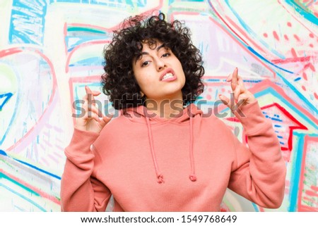 young pretty afro woman crossing fingers anxiously and hoping for good luck with a worried look against graffiti wall