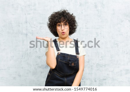 young pretty afro woman looking surprised and shocked, with jaw dropped holding an object with an open hand on the side