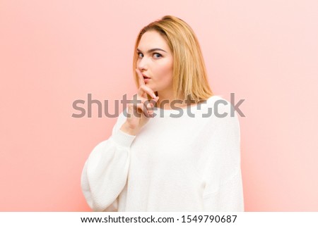 young pretty blonde woman asking for silence and quiet, gesturing with finger in front of mouth, saying shh or keeping a secret against pink flat wall
