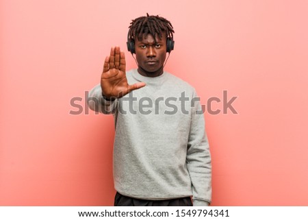 Young rasta black man listening to music with headphones standing with outstretched hand showing stop sign, preventing you.