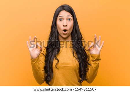 young pretty hispanic woman feeling shocked, amazed and surprised, showing approval making okay sign with both hands against brown wall