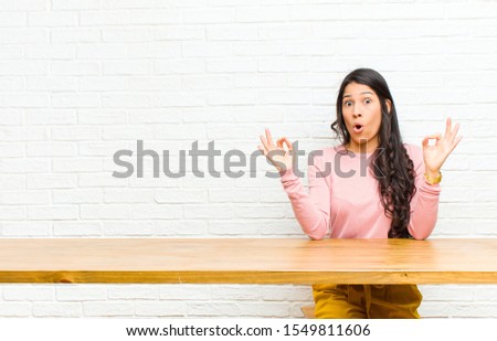 young  pretty latin woman feeling shocked, amazed and surprised, showing approval making okay sign with both hands sitting in front of a table