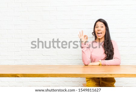 young  pretty latin woman feeling successful and satisfied, smiling with mouth wide open, making okay sign with hand sitting in front of a table