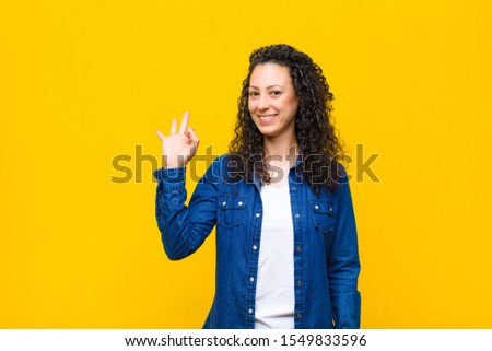 young pretty woman feeling happy, relaxed and satisfied, showing approval with okay gesture, smiling against orange wall