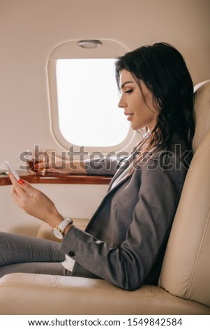 side view of businesswoman in formal wear holding glass with whiskey and smartphone in private jet 
