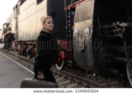 A girl with a vintage hair and a suitcase stands near an old locomotive of a train