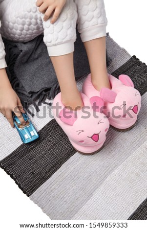 Cropped medium shot of a child in white textured pyjamas and pink plush house slippers made in the form of kawaii mouse. The child with a blue car is sitting on the striped carpet and a gray plaid.