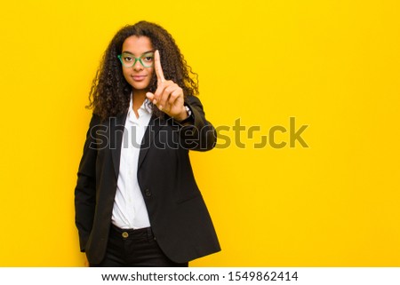 black business woman smiling proudly and confidently making number one pose triumphantly, feeling like a leader against orange wall