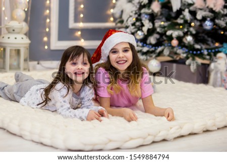 beautiful sister girls in white, gray and pink pajamas are lying on the carpet and smiling. a red cap on his head. In the background a Christmas tree, a garland, balls, gifts. horizontal photo.