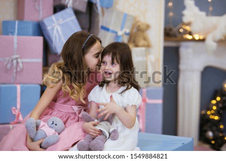 Emotion of surprise baby. Beautiful little girl with blond hair in a pink and white dress tells a secret in her ear. in the background are gift boxes of blue, purple, beige