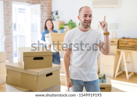 Young couple arround cardboard boxes moving to a new house, bald man standing at home pointing finger up with successful idea. Exited and happy. Number one.
